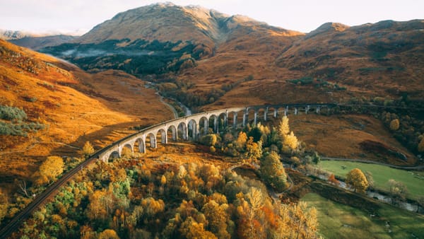 An aerial view of the historic, concrete Glenfinnan viaduct amid autumn-coloured foliage in a hilly landscape.