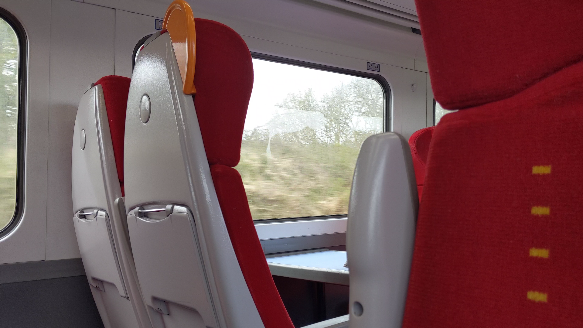There are seats with red moquette on the front and grey plastic on the back. On the backs of seats are fold-down tables, which are large enough to hold a drink, a snack, and a book. There are orange grab handles on the seats next to the walkways. There are grey plastic seat rests that can be folded down or kept up.