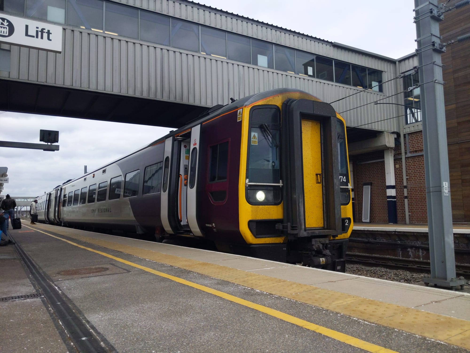 A Class 158 train is stationary at a station. The front of the train is painted vivid yellow and has two windows on either side separated by a protruding cab door. The livery is purple at the front. The doors are open and are white. The train is below a grey footbridge.