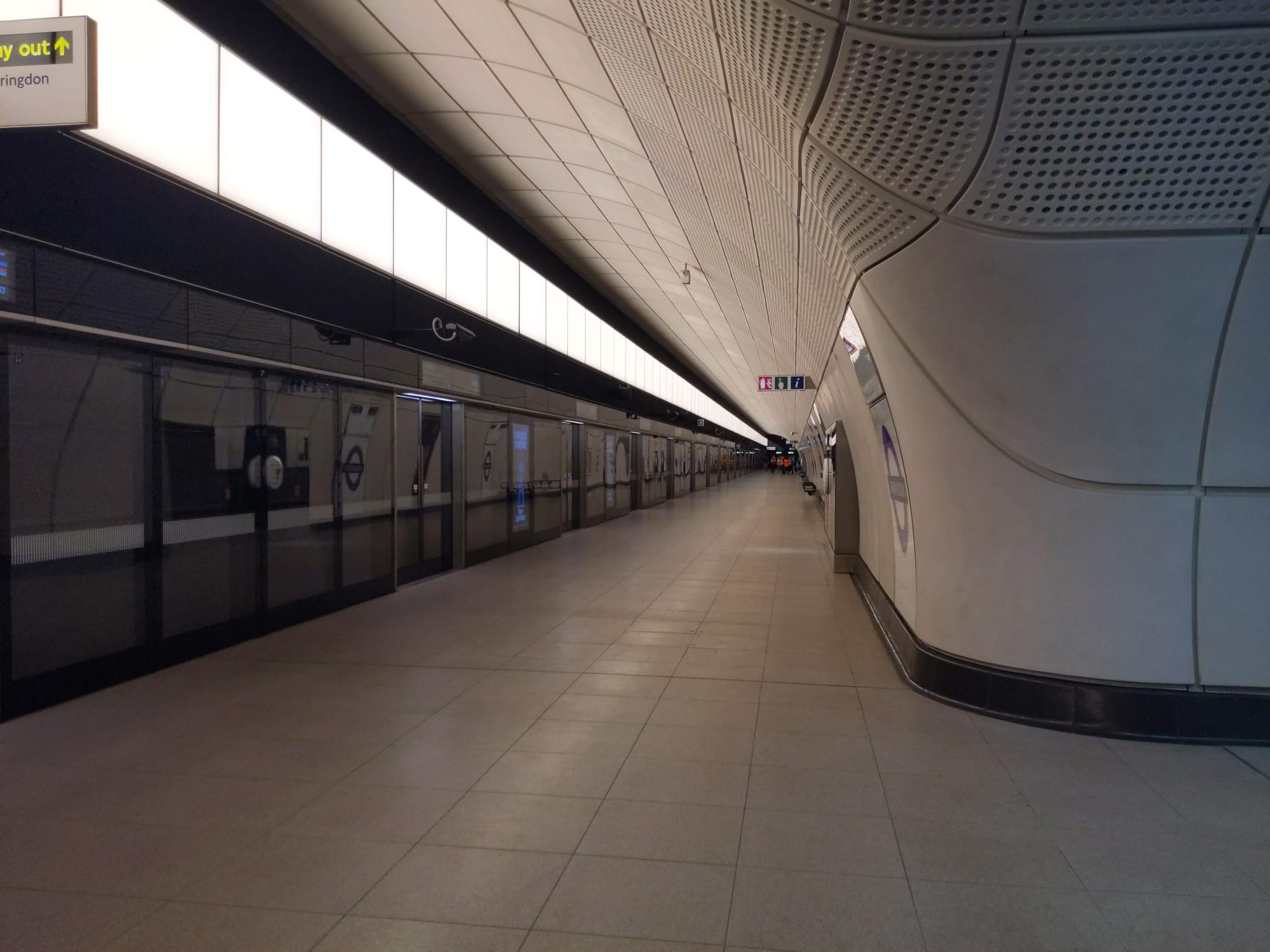 On the left are tinted platform screen doors. The wall extends to the ceiling of the arched platform and has a row of wide LED panels along the top. In the centre is the rest of the platform, which is several metres wide. The end of the platform in the background can hardly be seen.