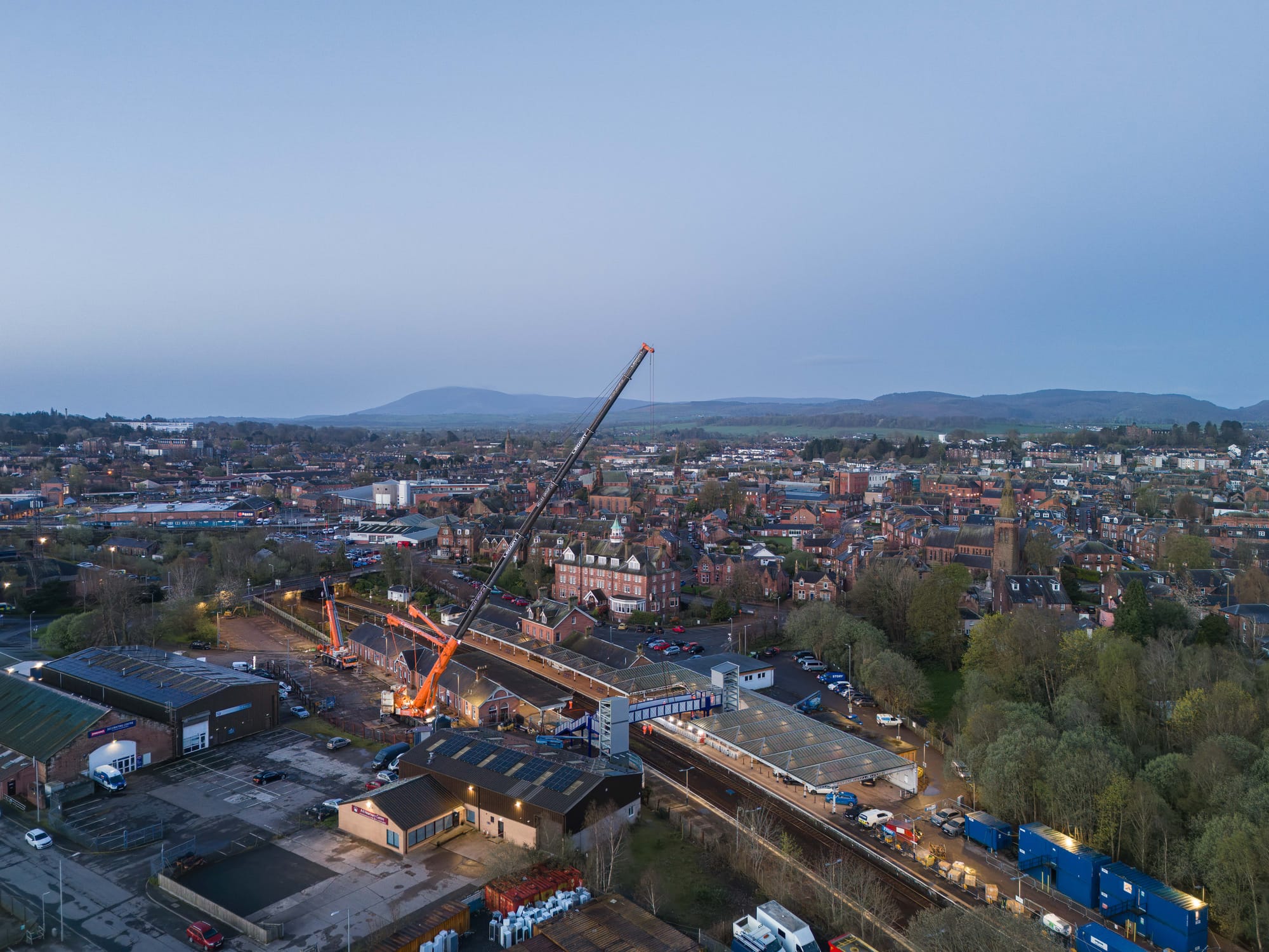 In the centre of the aerial image an orange and grey crane is seen lowering the footbridge into place. The buildings of the town can be seen behind the station and crane. In the far distance the hilly landscape can be seen.