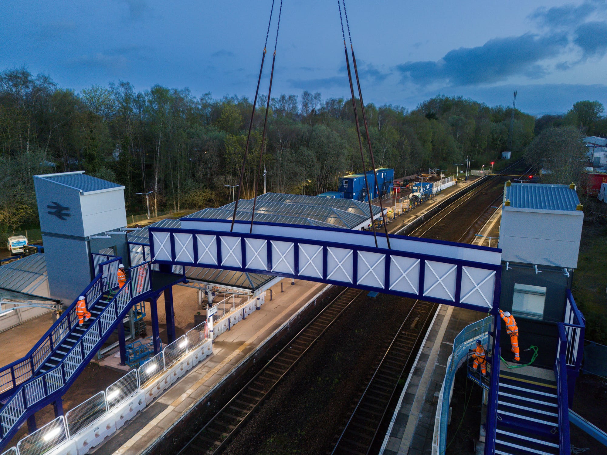 The white and blue footbridge, as seen from above, is lowered by a crane (out of frame) between two sets of stairs.