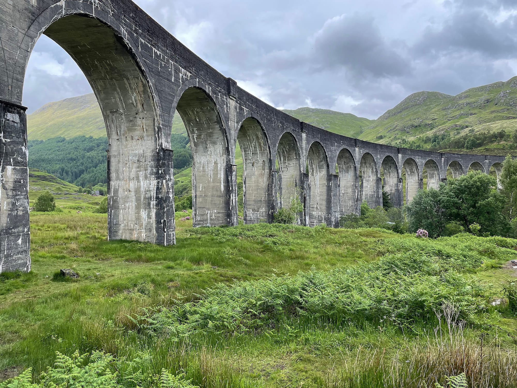 A view from the ground of Glenfinnan viaduct with multiple arches stretching across a lush green landscape with hills in the background on an overcast day.
