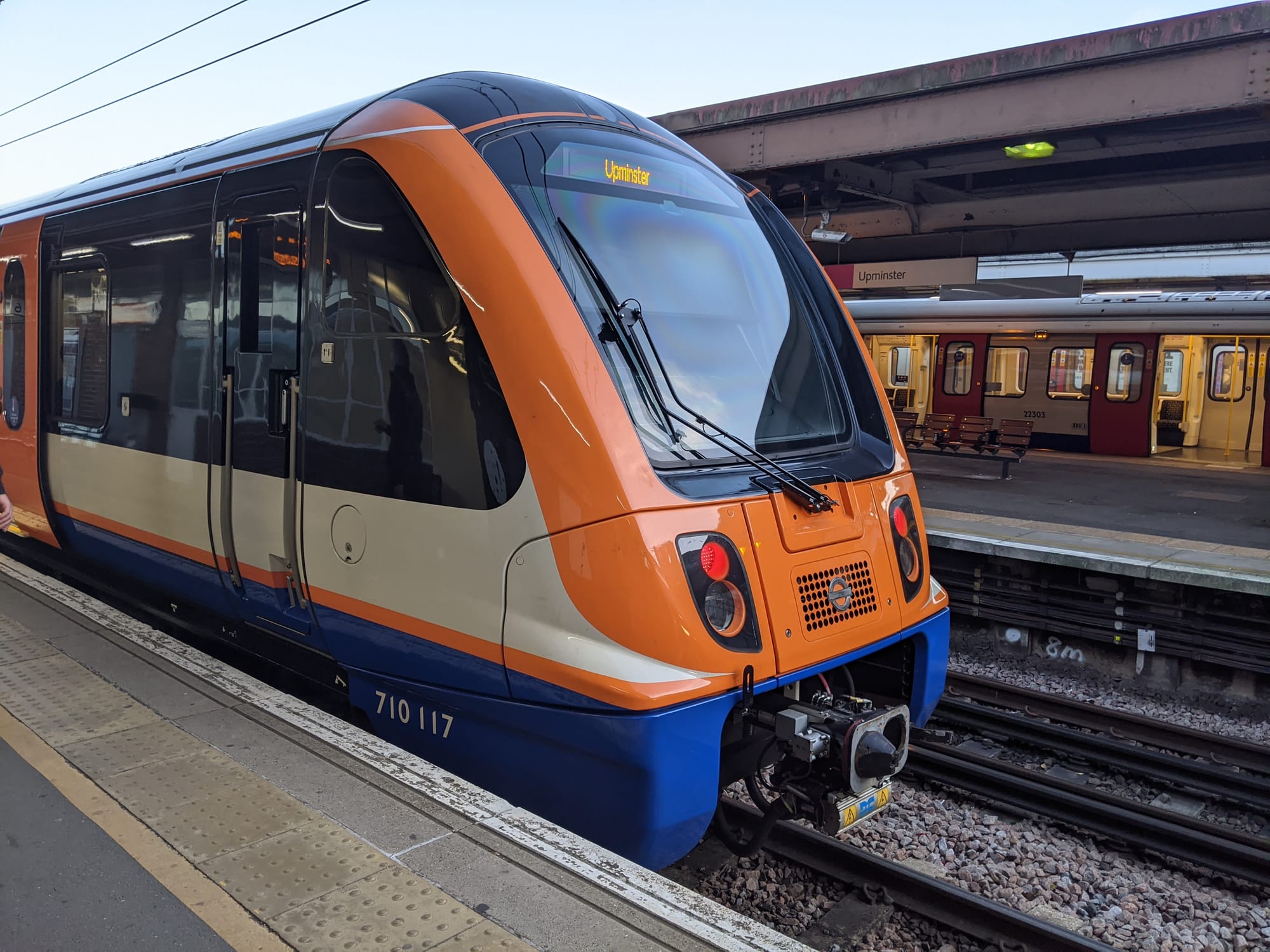 A London Overground train is stationary at a platform. The red rear lights are illuminated on the back of the train. The back of the train is painted bright orange around the outside of a large window. The bottom part of the train is painted blue.