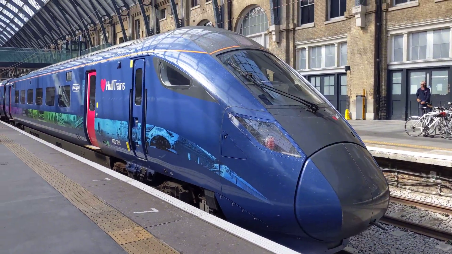 The train is in a Hull Trains livery, which is black on the pointed front of the train. Along the side of the train is a deep blue with a composite image of landmarks around Hull. The doors slide into the side of the train and are bright pink.
