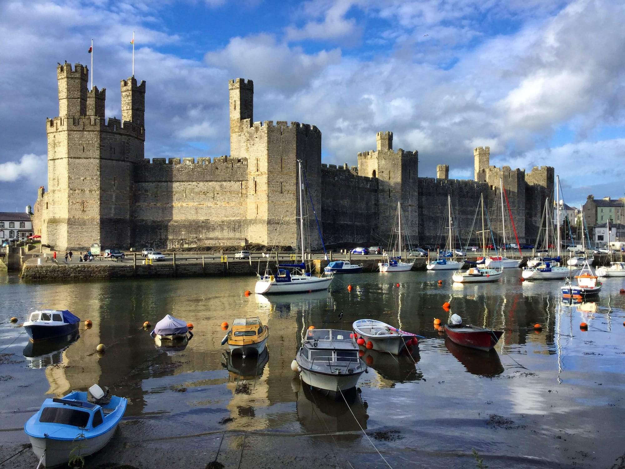 A view of Caernarfon Castle in Wales, with its massive stone walls and prominent towers. The medieval fortress is situated by a harbour filled with various small boats, under a partly cloudy sky.