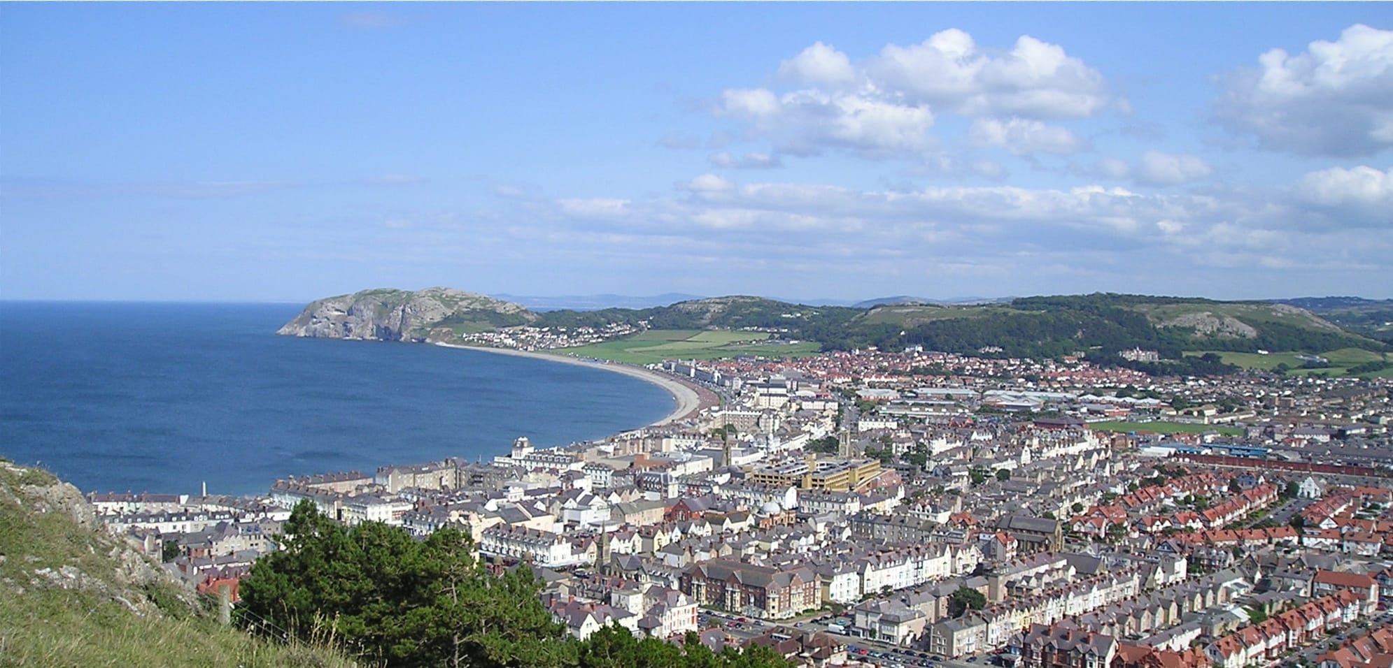 A high-up view of Llandudno, a coastal town in Wales. The town's buildings and rooftops stretch along the shoreline, curving round a wide bay with a sandy beach. The hills and headland of Little Orme can be seen in the background.