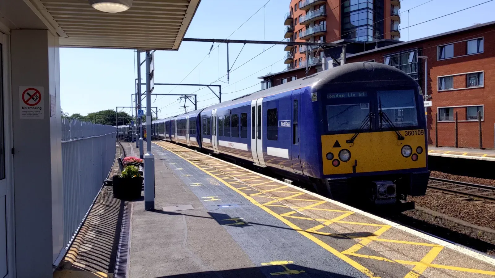 A class 360 train departs Witham with its blue livery and white doors. The rear of the train is painted yellow. It is a summer day with a clear, blue sky.