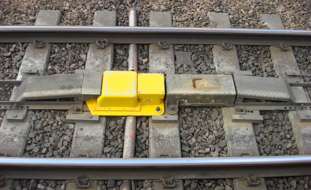 An AWS magnet is pictured in the middle of some tracks with a metallic permanent magnet and bright yellow electromagnet. It is fixed atop the concrete sleepers and is surrounded by ballast.