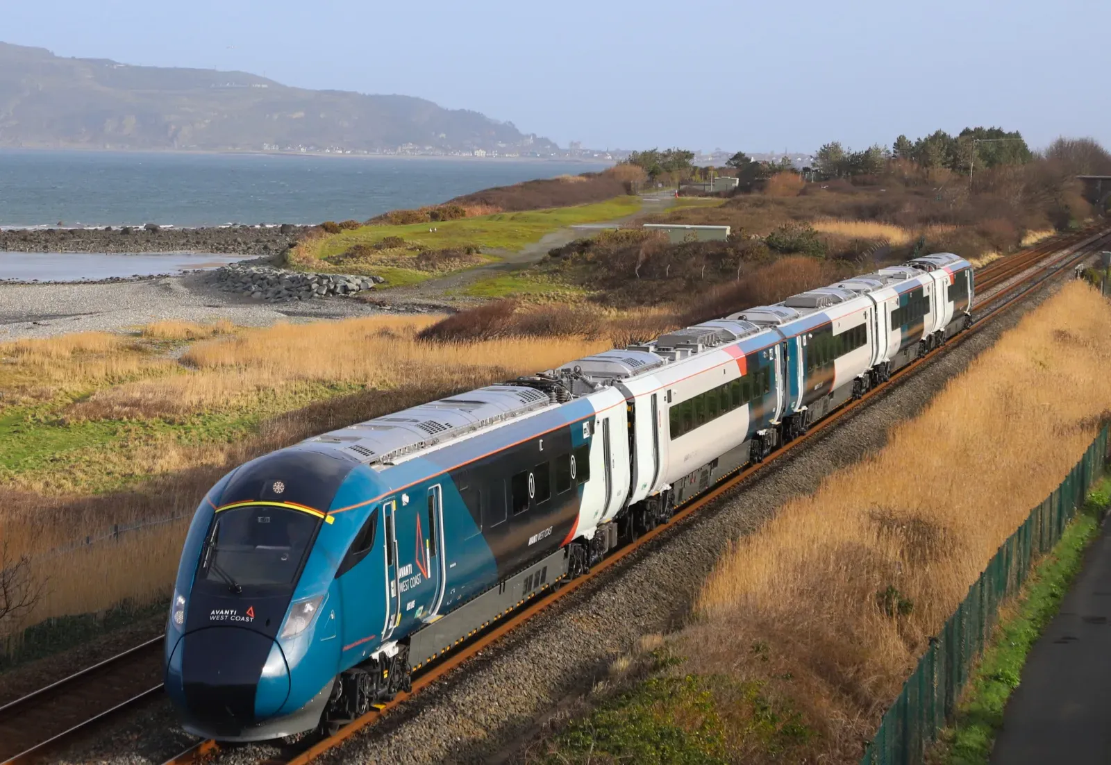 A class 805 Evero is travelling down a coastal railway track with its teal and white livery. In the near distance is a rocky shoreline. Beyond the stretch of water are distant hills under a clear, blue sky.