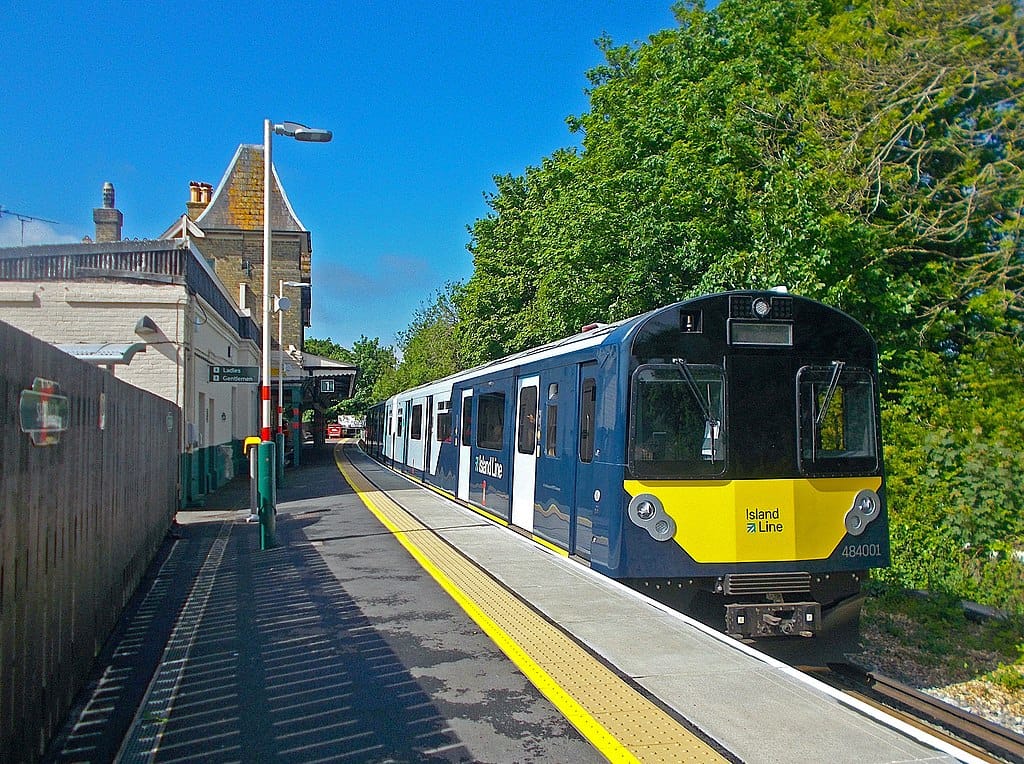 A train with a deep blue and white livery sits stationery alongside a platform. It is a single-track station. To the right of the train is dense foliage, and there is a clear blue sky up above.