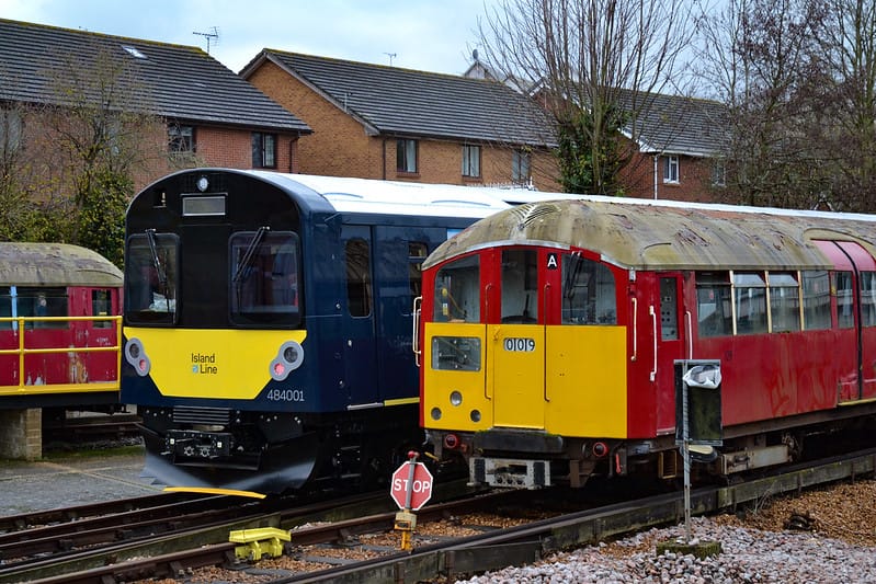 A taller, modern train with a deep blue livery is alongside its predecessor in a red livery. Both trains are in the depot.