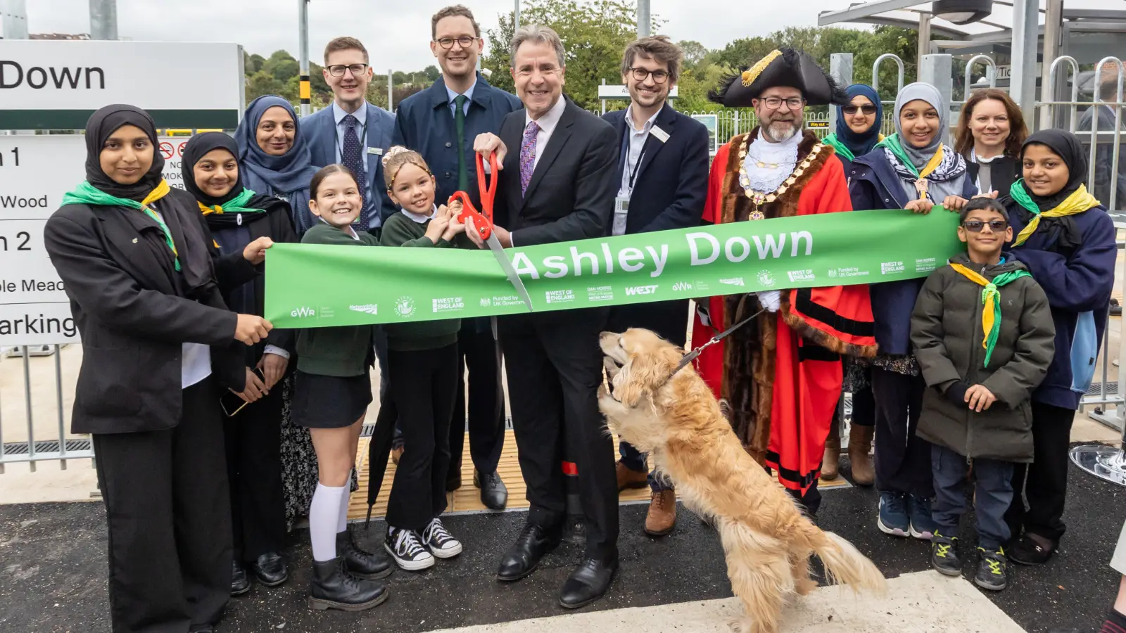 A crowd of people surround a lime green banner which reads 'Ashley Down' in bold, white font. A comically large pair of scissors is being used to cut the banner to signify the station's opening.