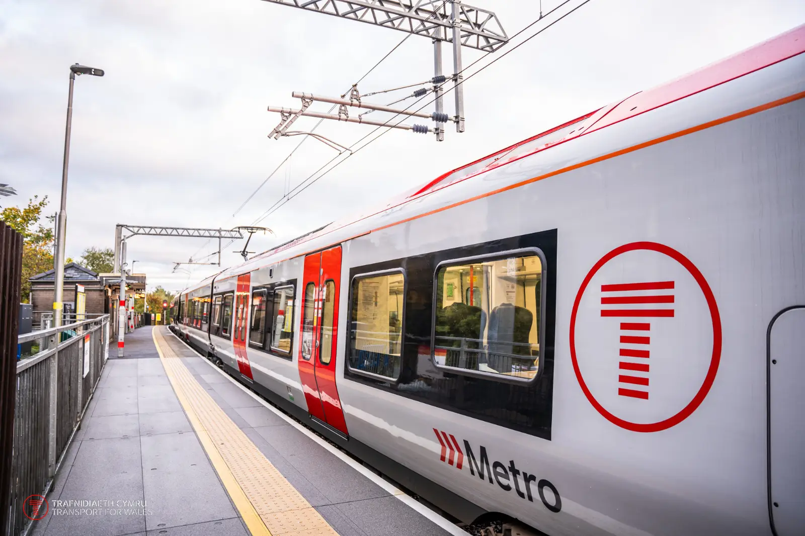 Modern train labeled with the Transport for Wales (Trafnidiaeth Cymru) branding, featuring a red 'T' logo inside a circle and the word 'Metro' on its side. The train is stationed at a platform under overhead electric lines, with a clean and modern railway station in the background.