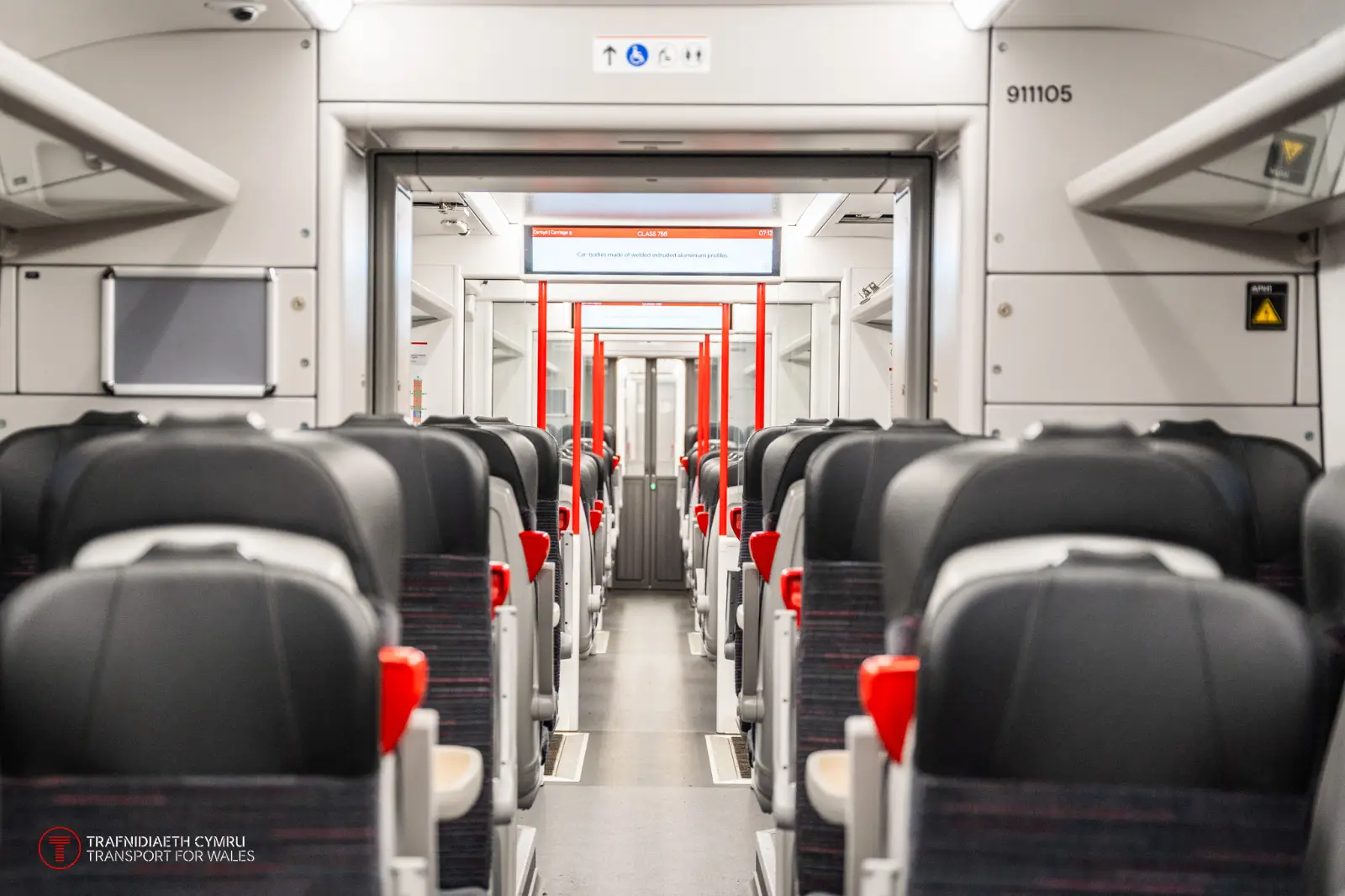 Interior view of a modern Transport for Wales (Trafnidiaeth Cymru) train, showing rows of black and red seats with armrests and headrests. The aisle is clean and spacious, with overhead luggage racks and clear signage displayed on electronic screens above. Red poles and sleek design elements enhance the contemporary appearance of the train carriage.