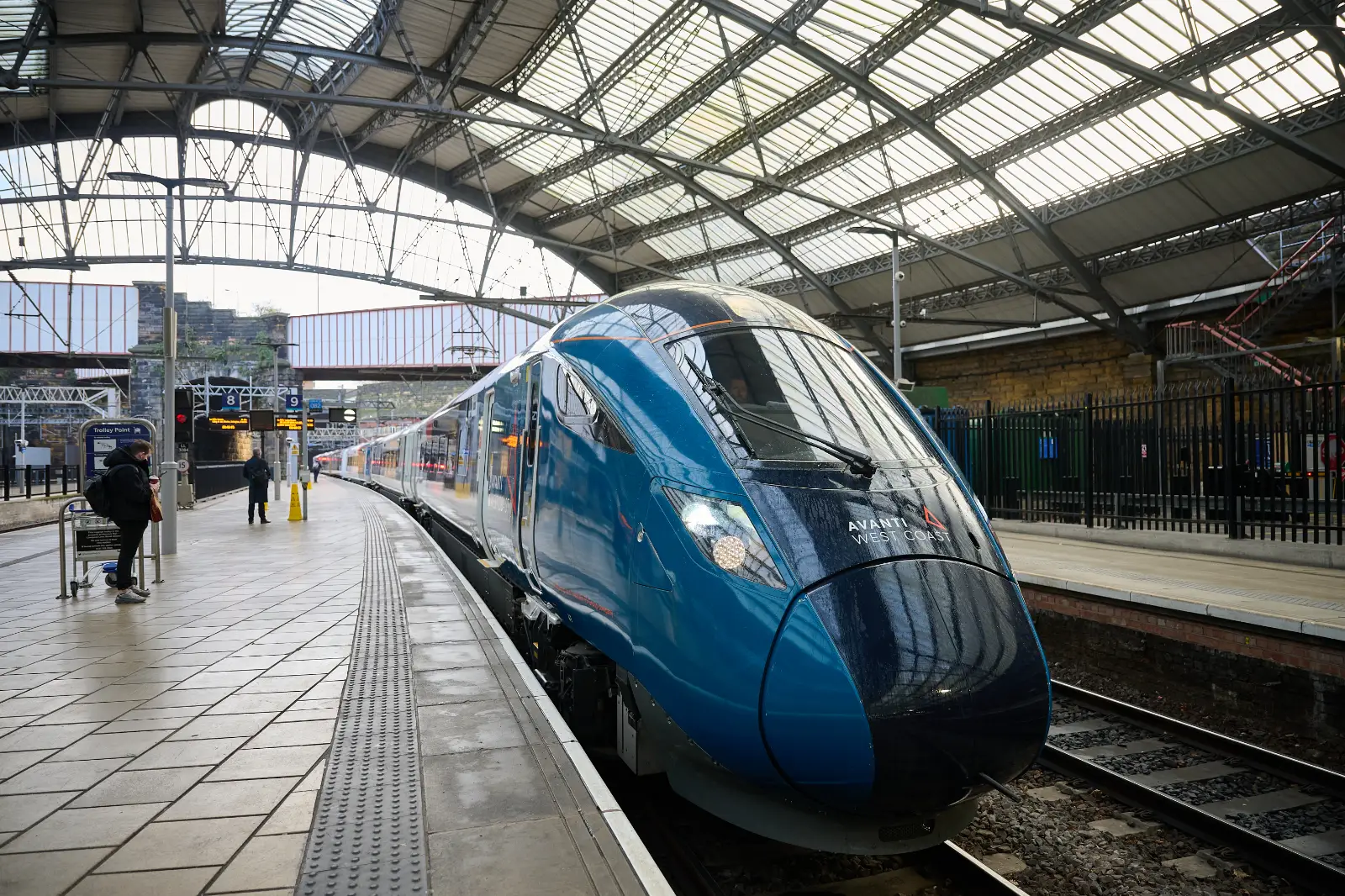 The image shows an Avanti West Coast train at a station platform under a large, arched glass and metal roof. The train, in a sleek dark blue livery, is facing the viewer and positioned along a curved platform. A few people are waiting nearby, with one person using a luggage trolley. Digital signs display platform numbers and information in the background. The station has a historic, industrial architecture with a high roof and metal beams.