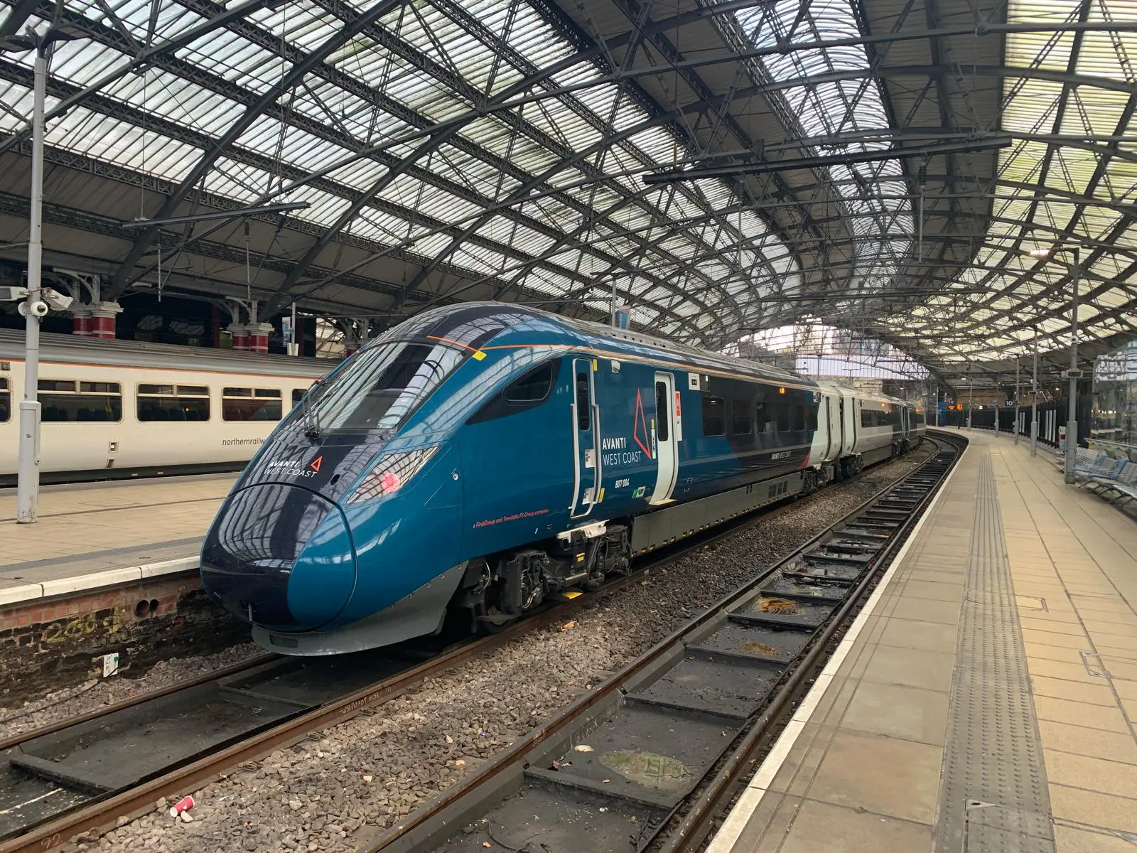 The image shows an Avanti West Coast train at a station platform, with the train positioned on the curved track under an arched, glass-roofed structure. The train is in a dark blue livery with the Avanti West Coast logo. The platform is empty, with seating along the side, and another train can be seen in the background on a parallel track.