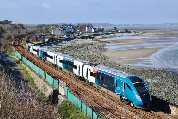 The class 805 travels along a costal railway track under a clear sky. It has a teal and white livery with accents of orange.