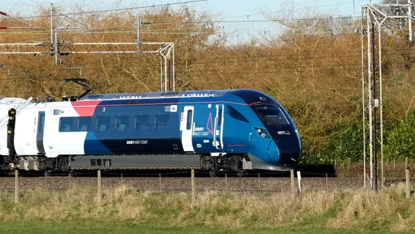 The Class 805 moves at speed along a single-track railway, under overhead lines. There is lots of foliage in the background.