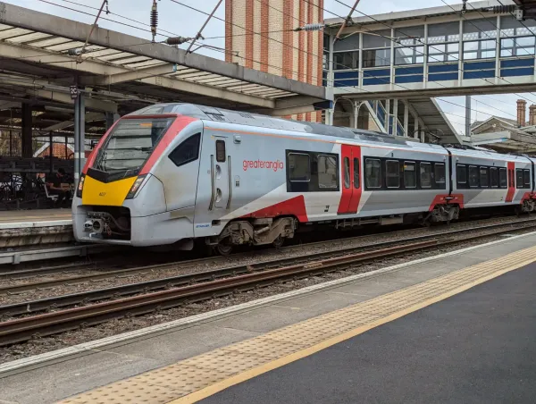 A Greater Anglia train with a silver livery is stationary beside a platform. It has a bright yellow front, with red accents.