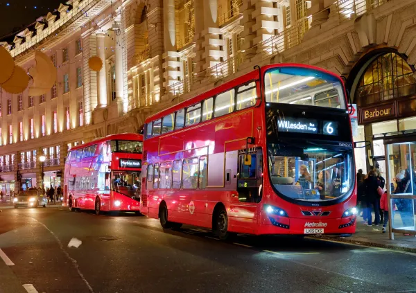 Two London buses in their distinctive, bright red livery line up at a bus stop in the city.