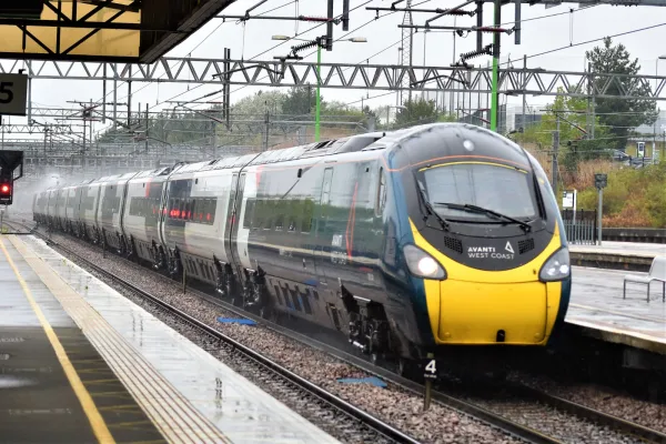 An Avanti West Coast train speeds through a station in the rain. It has a yellow front, turquoise livery with orange accents.