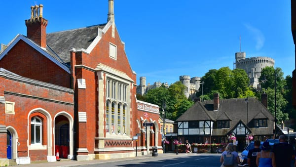 The front of the red-brick Windsor & Eton Riverside station with a cottage to the right and Windsor Castle in the background.